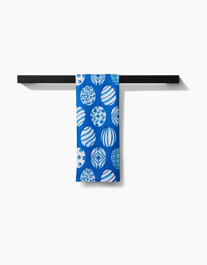 A Delft Blue Easter Eggs tea towel by Sarah Daugherty, featuring white and blue circular designs and made from recycled materials, hangs over a black towel bar against a white background.