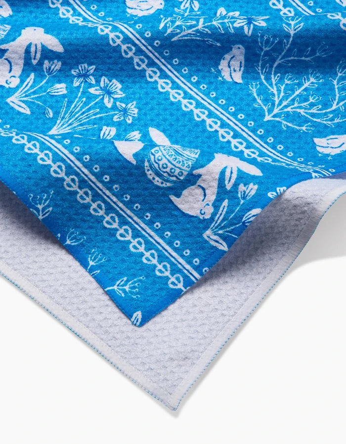 A close-up of the Sarah Daugherty Delft Blue Easter Stripe tea towel, featuring white rabbits, flowers, and decorative patterns on blue, layered over light grey textured fabric.