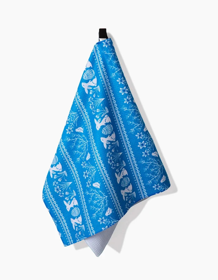 The Sarah Daugherty Delft Blue Easter Stripe recycled kitchen towel with white fish, coral, and floral patterns hangs from a black loop against a plain white background.
