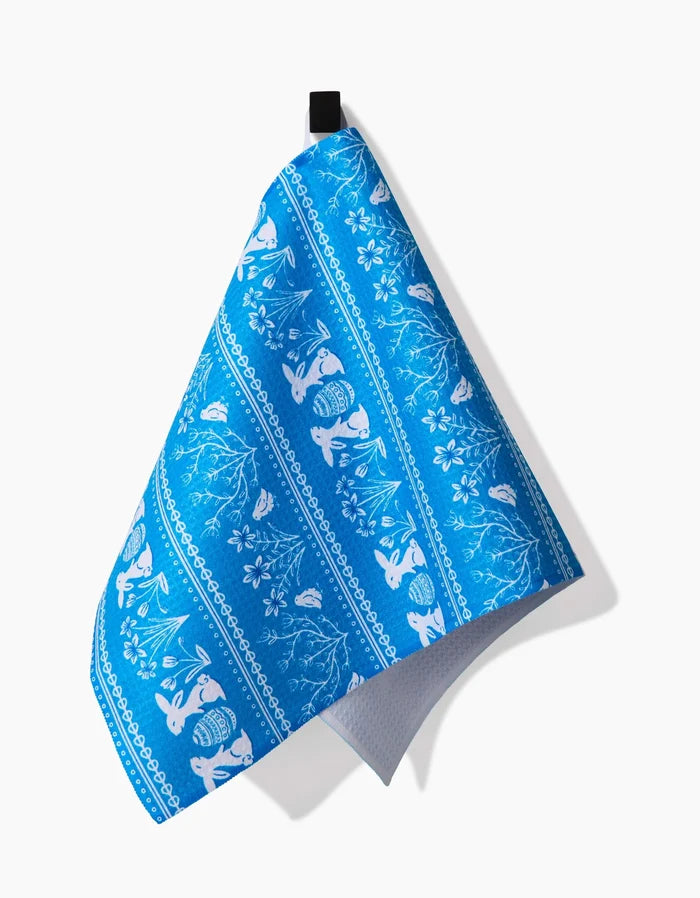 The Delft Blue Easter Stripe bar towel by Sarah Daugherty features a blue and white pattern with rabbits, flowers, and leaves, made from recycled materials and hanging by a black loop.