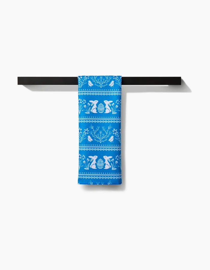 The Delft Blue Easter Stripe kitchen towel by Sarah Daugherty, featuring white animal and plant patterns, is draped over a black towel bar against a plain white background.