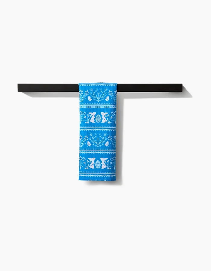 The Delft Blue Easter Stripe bar towel by Sarah Daugherty, featuring white bird and tulip patterns on blue Geoweave fabric, hangs neatly on a black bar, adding charm and function against a crisp white backdrop.