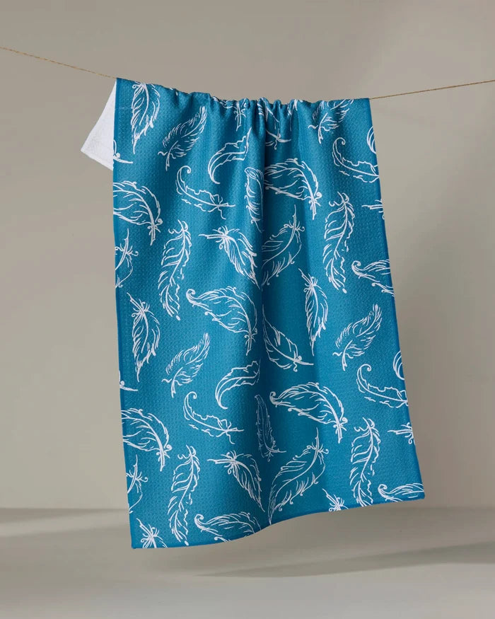 The Elena Fay “Delicate Feathers” tea towel, quick-drying and crafted from recycled materials, features a blue color with a white feather pattern and hangs on a clothesline against a neutral backdrop.