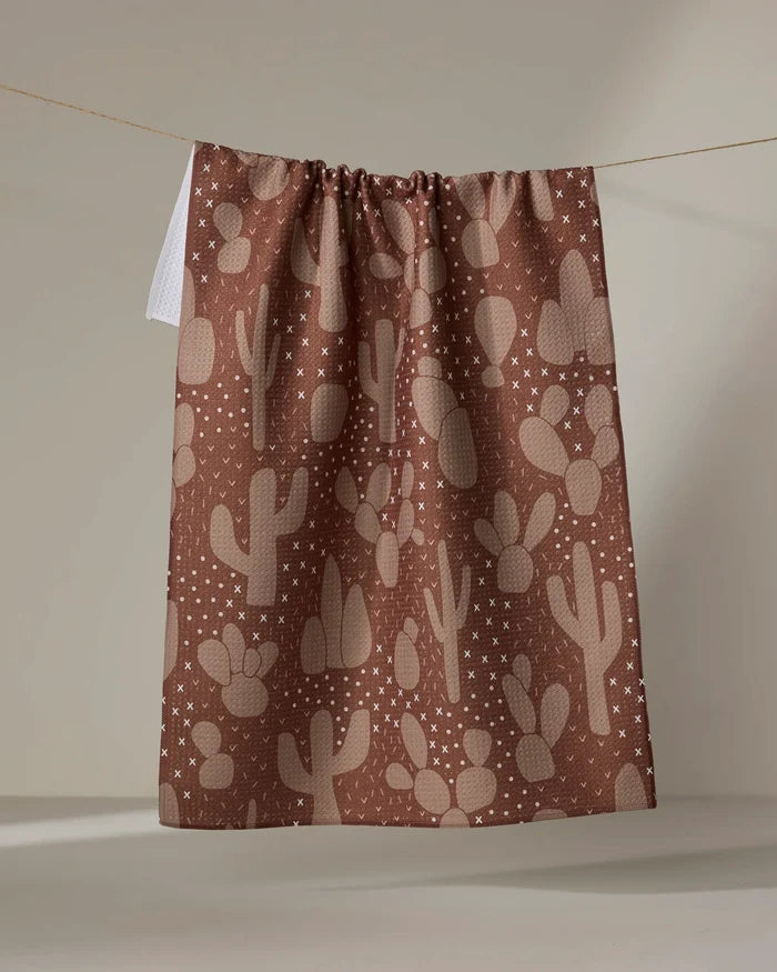 The "Desert Night" tea towel by Rosa Kichline, featuring a beige cactus and star pattern on brown, hangs on a line against a neutral background.