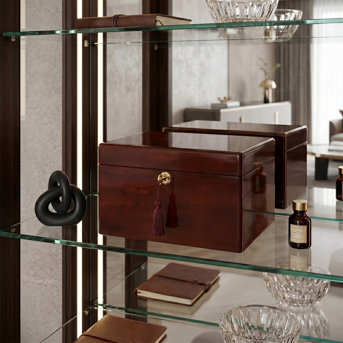 Brown urn box with a gold clasp on a glass shelf with decorative items.
