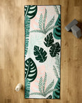 The GEOMETRY Diedre yoga mat with a green and pink tropical leaf pattern lies on a wooden floor, topped with a non-slip yoga towel. Nearby are two stacked yoga blocks and a water bottle.
