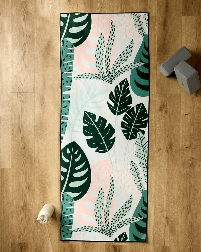 The GEOMETRY Diedre yoga mat with a green and pink tropical leaf pattern lies on a wooden floor, topped with a non-slip yoga towel. Nearby are two stacked yoga blocks and a water bottle.