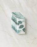 Five neatly stacked GEOMETRY Diedre recycled paper towels, featuring green tropical leaf patterns, are displayed on a light marble surface with subtle veining.