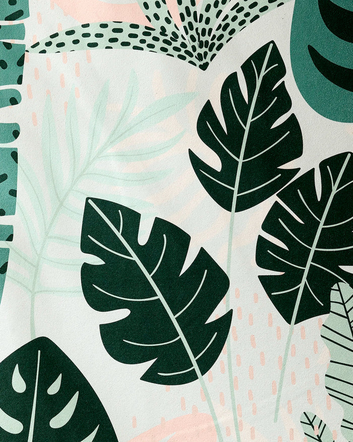 The Diedre yoga towel by GEOMETRY features abstract botanical art with large dark green monstera leaves, pale green ferns, and soft pink accents on a light background for a modern tropical look and non-slip performance.