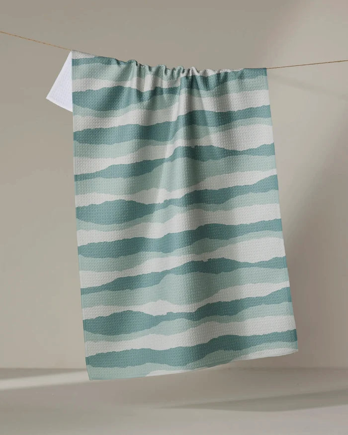 The Distant Landscape tea towel by Twig + Spark, featuring quick-dry fabric and teal and white wavy horizontal stripes, hangs on a clothesline against a neutral backdrop.