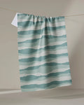 The Distant Landscape tea towel by Twig + Spark, featuring quick-dry fabric and teal and white wavy horizontal stripes, hangs on a clothesline against a neutral backdrop.