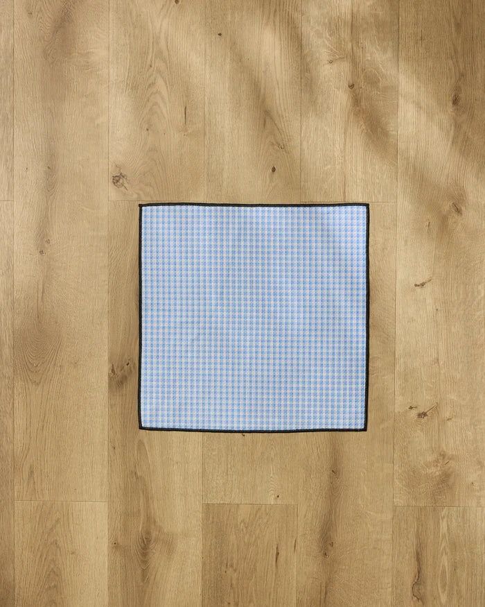 The Geometry Dog Delight towel, featuring blue and white checks, a black border, and ultra-absorbent material, is laid flat on a light wood floor.