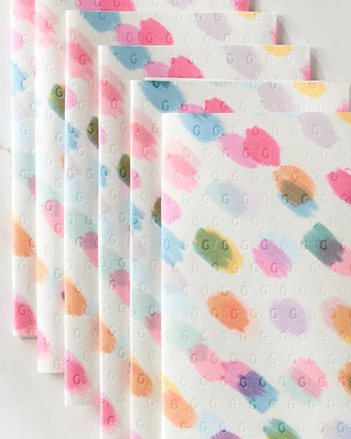 Six stacked, reusable Downpour towels by GEOMETRY feature pastel swatches in pink, blue, orange, green, and purple. Each white eco-friendly napkin is embossed all over with small repeating “G” letters.