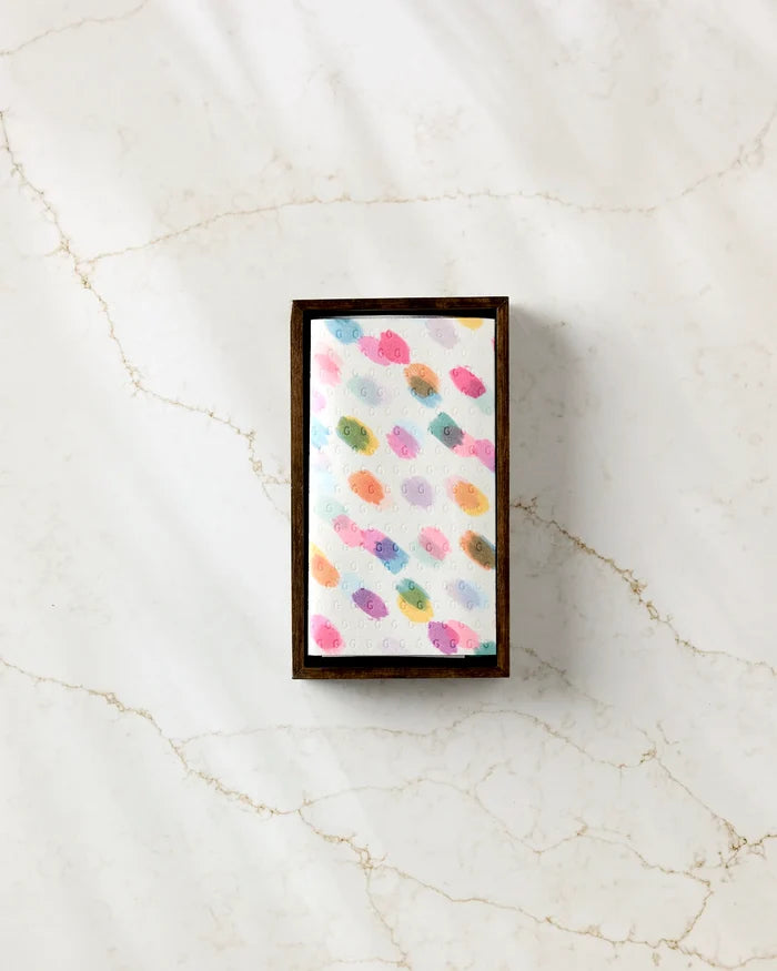 The GEOMETRY Downpour soap, a rectangular white bar with colorful fish designs, rests in a dark wooden dish on veined marble beside a stack of eco-friendly paper towels.