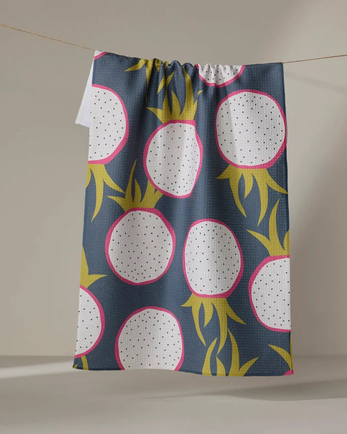 The Lizzie Clark "Dragon Fruit Frolic" tea towel, featuring bold dragon fruit shapes in pink, white, yellow-green, and black on dark blue fabric, dries quickly and hangs on a line.