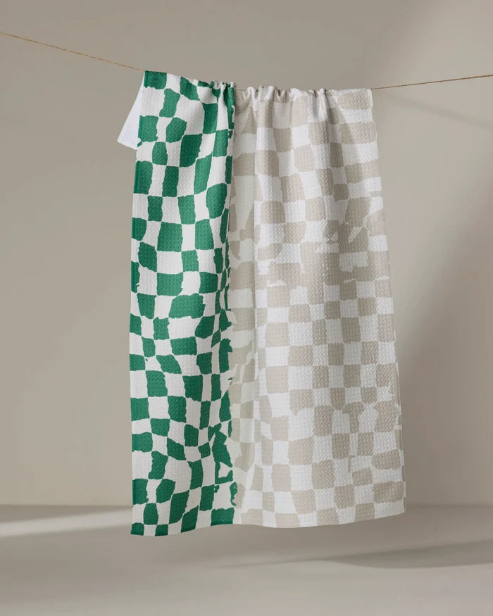 The Dual tea towel by Geometry, made from recycled materials, hangs on a neutral background. Its left side displays green and white uneven checks, while the right side features beige and white checks in a checkered pattern.