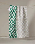 The Dual tea towel by Geometry, made from recycled materials, hangs on a neutral background. Its left side displays green and white uneven checks, while the right side features beige and white checks in a checkered pattern.