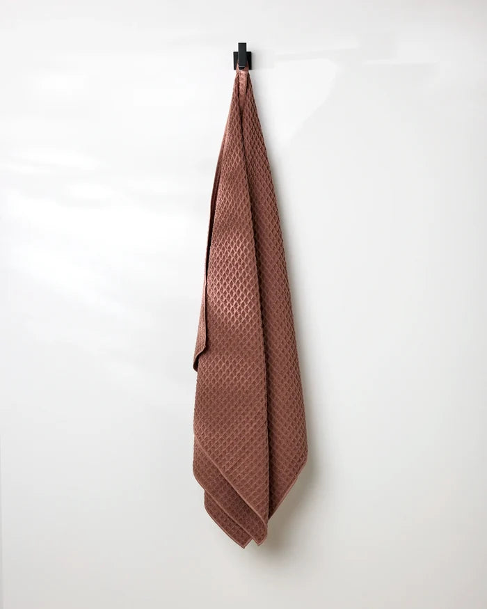 A GEOMETRY Dusty Rose Waffle Bath Towel hangs neatly from a black hook on a plain white wall.