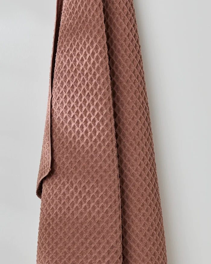 The GEOMETRY Dusty Rose waffle bath towel, with its textured rose hue, hangs against a plain light gray wall, bringing understated luxury to your space.