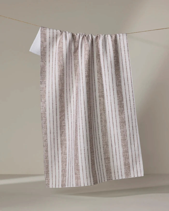 The Becki Owens Dyer Rust tea towel, featuring beige and white tones with vertical brown dotted stripes, hangs neatly over a rope against a neutral background.