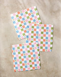 Three square napkins from Kayla Ann's Earth Day Checkers set, with vibrant checks and icons like hearts, stars, and flowers, are displayed on a beige surface—ideal as kitchen towels or recycled dishcloths.