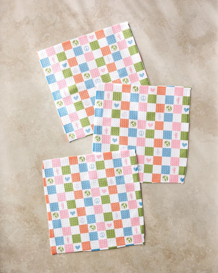 Three square napkins from Kayla Ann's Earth Day Checkers set, with vibrant checks and icons like hearts, stars, and flowers, are displayed on a beige surface—ideal as kitchen towels or recycled dishcloths.