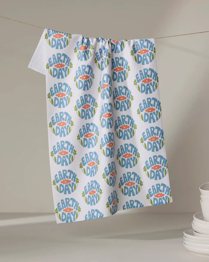 A white, ultra-absorbent Kayla Ann Earth Day Stamp tea towel hangs on a line, adorned with blue, green, and orange circular patterns. Stacks of white bowls and plates are arranged on the right side of the table below.