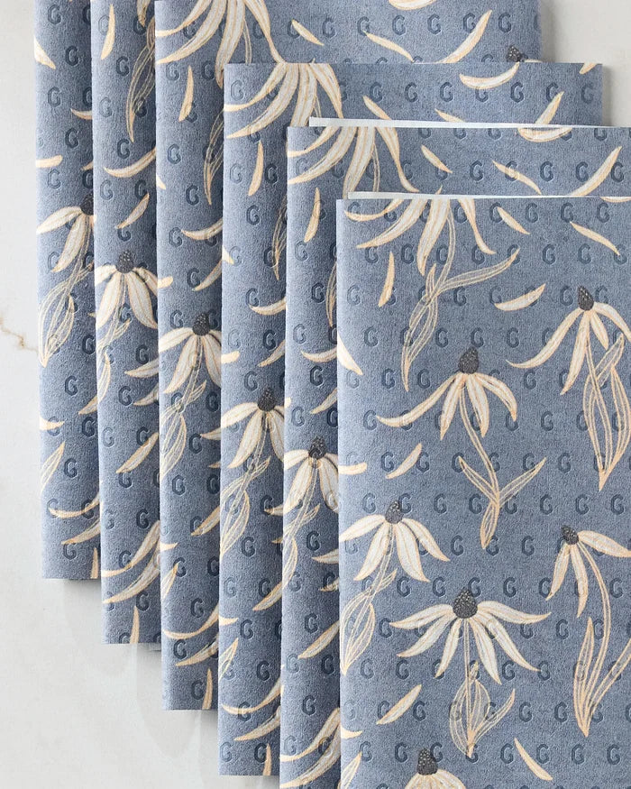 Six stacked Echinacea napkins by Katie O'Shea, featuring a beige and white floral design with blue G letters, displayed fan-style on marble—an elegant, eco-friendly paper towel choice for any occasion.