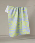The Electric Tiger tea towel by Elisabeth Wing, featuring light purple and yellow zebra stripes and made from recycled materials, hangs on a clothesline against a neutral backdrop.