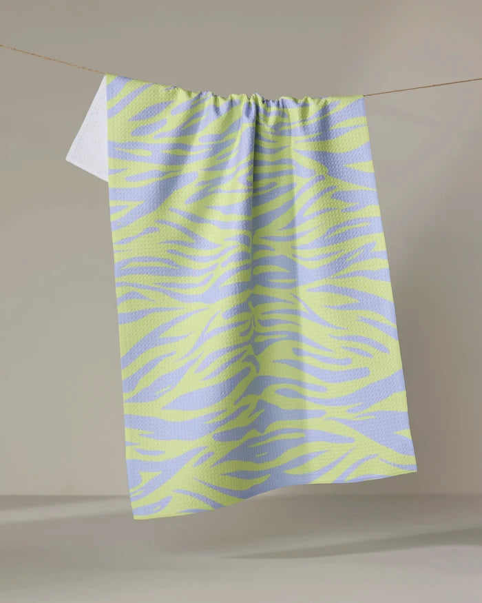 The Electric Tiger tea towel by Elisabeth Wing, featuring light purple and yellow zebra stripes and made from recycled materials, hangs on a clothesline against a neutral backdrop.