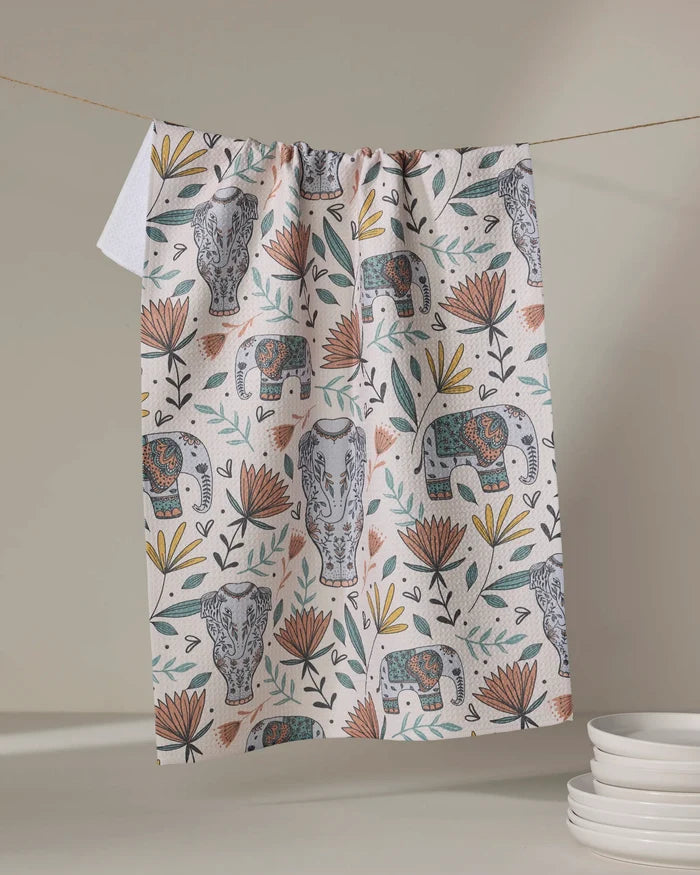The Amita Nair "Elephant Jungle" kitchen towel, decorated with elephants, big cat faces, and flowers in earthy hues, hangs on a string above stacked white plates on the right.