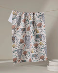 The Amita Nair "Elephant Jungle" kitchen towel, decorated with elephants, big cat faces, and flowers in earthy hues, hangs on a string above stacked white plates on the right.
