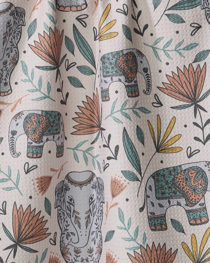 The "Elephant Jungle" tea towel by Amita Nair features decorated teal and orange elephants with green, yellow, and orange leaves and flowers on a light background. Made from recycled materials for sustainability.