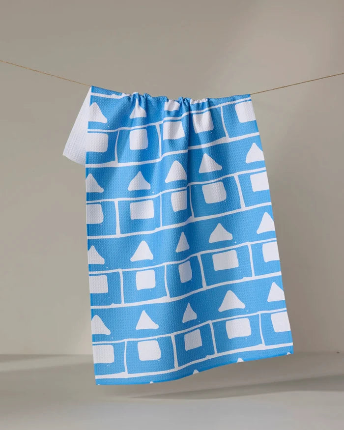 The Sissy Moon "Elevation" tea towel hangs on a line, showcasing quick-drying performance and a blue background with geometric white squares and triangles arranged in horizontal rows.