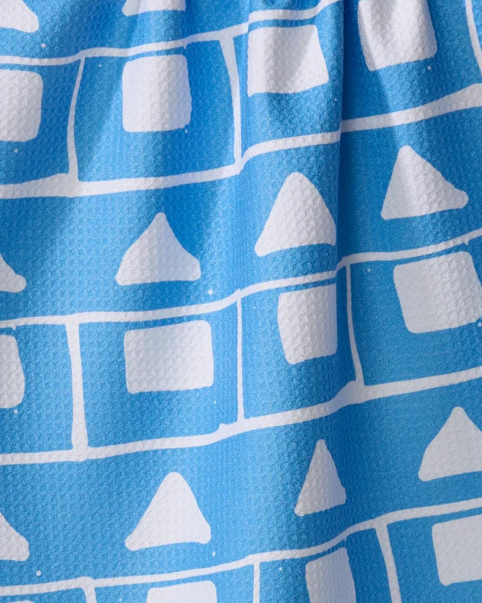 A close-up of Sissy Moon’s Elevation kitchen towel shows blue, textured recycled fabric with rows of white rectangles and triangles, featuring subtle folds and shadows.