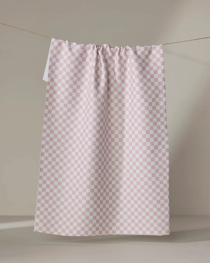The Ella tea towel by Charlie Rowan Designs, featuring a pale pink and white checkered pattern, hangs on a string against a plain beige background with soft lighting and gentle shadows.