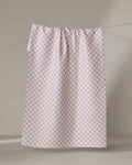The Ella tea towel by Charlie Rowan Designs, featuring a pale pink and white checkered pattern, hangs on a string against a plain beige background with soft lighting and gentle shadows.