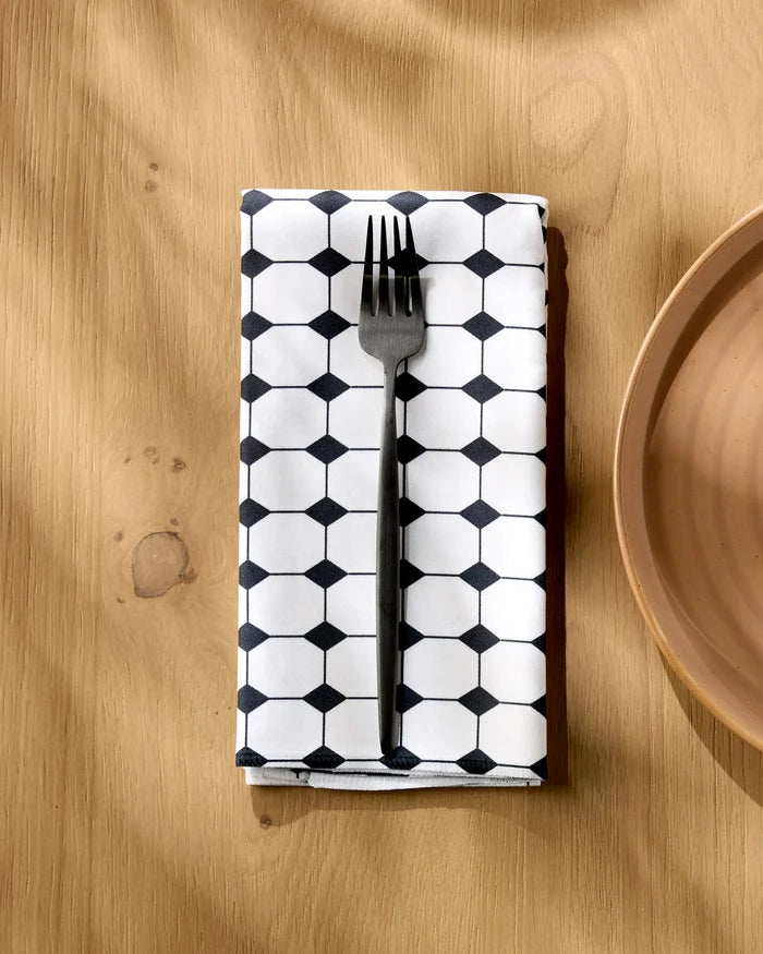 A metal fork rests on a Geometry Espresso Mosaic absorbent dinner napkin, featuring a black-and-white hexagon pattern, placed on a light wooden table beside a beige ceramic plate.