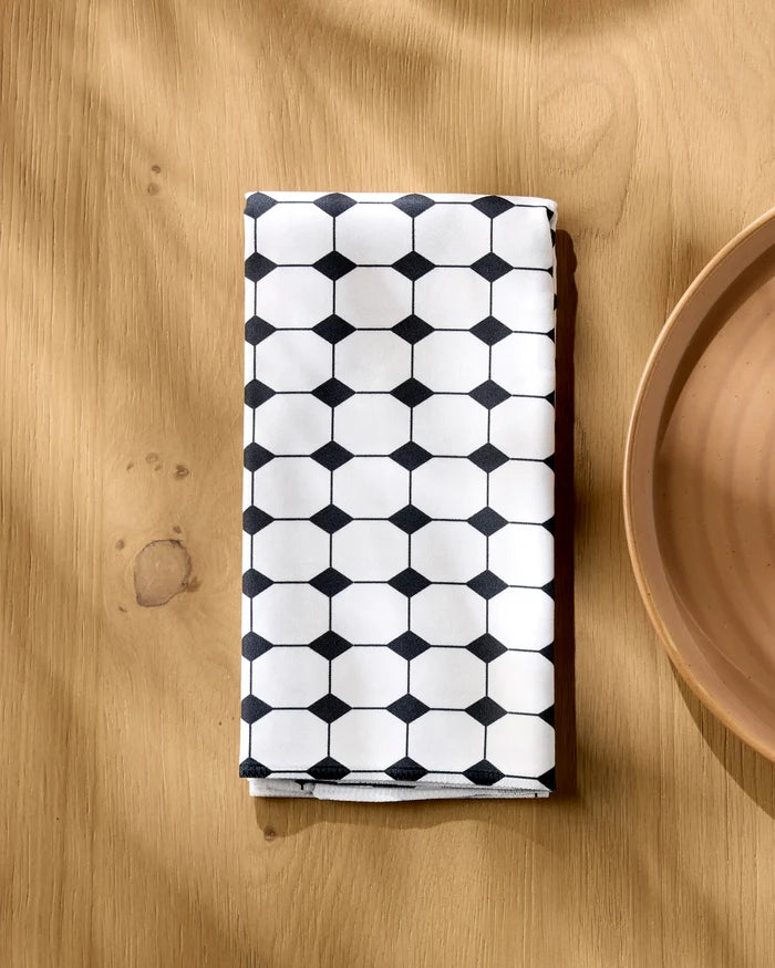 The Geometry Espresso Mosaic napkin, featuring a black hexagon and dot pattern on white, is folded neatly on a light wooden table beside the edge of a tan ceramic plate.