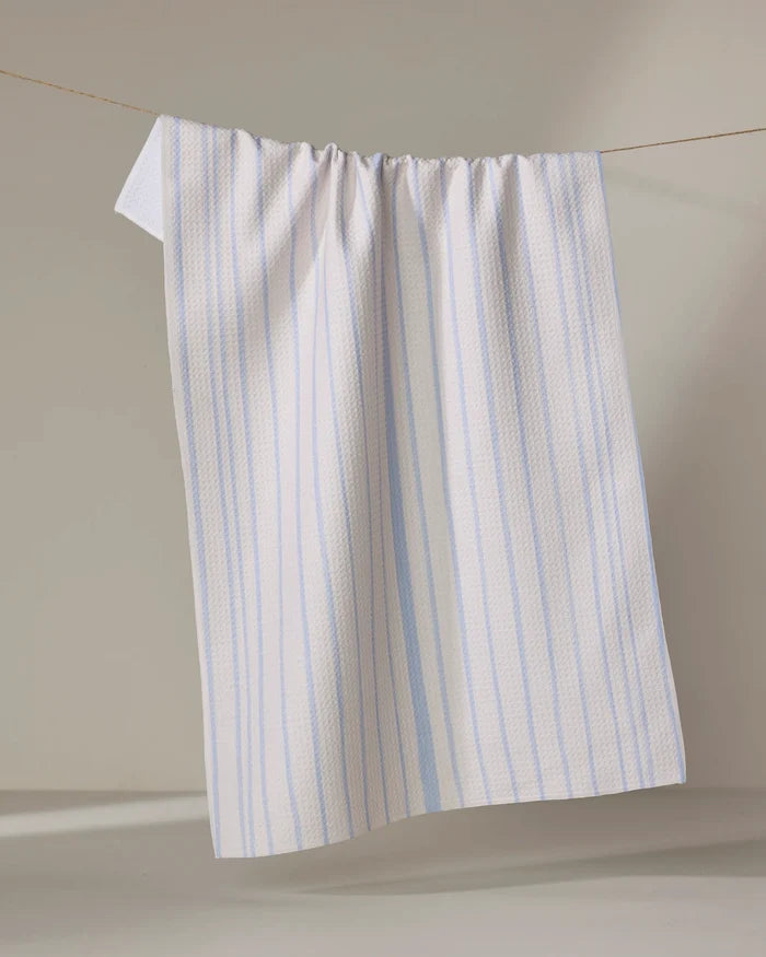 The Eve Stripe dish towel by Becki Owens, featuring white fabric with light blue vertical stripes, hangs on a clothesline against a neutral background.
