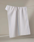 The Eve Stripe dish towel by Becki Owens, featuring white fabric with light blue vertical stripes, hangs on a clothesline against a neutral background.