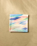 The Eventide square scarf by Rachel Parker features a pastel geometric pattern in blue, pink, green, and cream, neatly folded on a sandy beige surface reminiscent of a soft, quick-drying beach blanket.