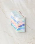 Seven Eventide recycled paper towels by Rachel Parker, each folded and featuring pastel diagonal stripes in blue, pink, and green, are neatly arranged on a light marble surface with beige veining.