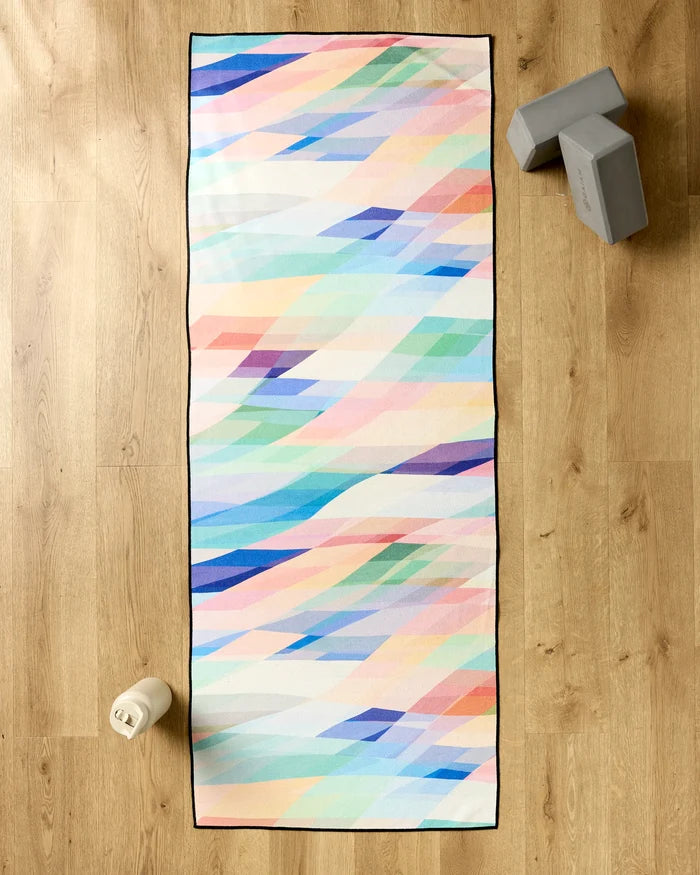 The Rachel Parker Eventide yoga mat, featuring a pastel geometric design, is spread on a wooden floor with a slip-resistant towel. Gray yoga blocks and a white water bottle are placed next to the mat.