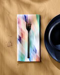 A metal fork rests on a folded Eventide recycled dinner napkin by Rachel Parker, beside a dark blue ceramic plate on a light wooden table. Sunlight casts gentle shadows over the eco-friendly surface.