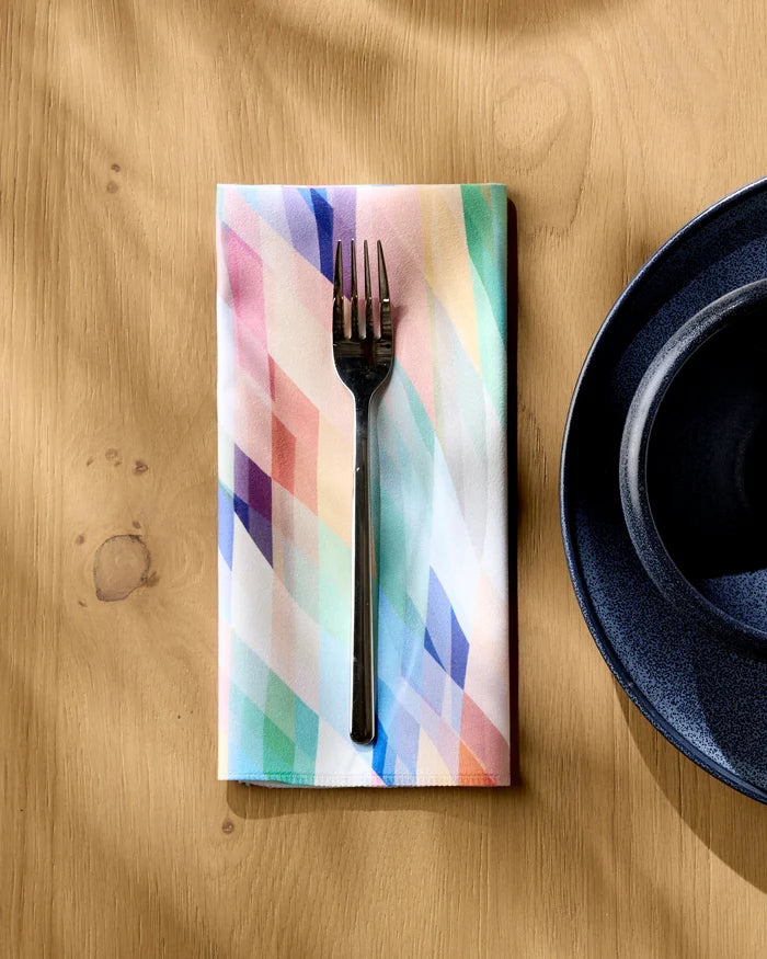 A metal fork rests on a folded Eventide recycled dinner napkin by Rachel Parker, beside a dark blue ceramic plate on a light wooden table. Sunlight casts gentle shadows over the eco-friendly surface.