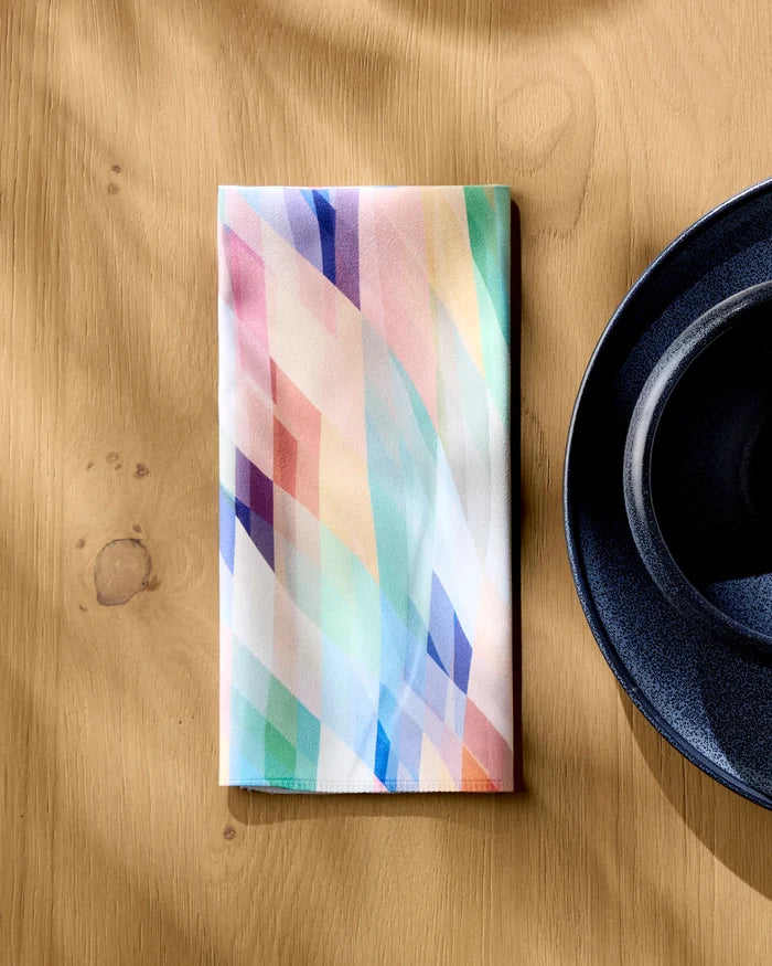 The Eventide napkin by Rachel Parker, featuring pastel abstract diagonal stripes and eco-friendly materials, is folded on a light wooden table next to a dark blue plate.