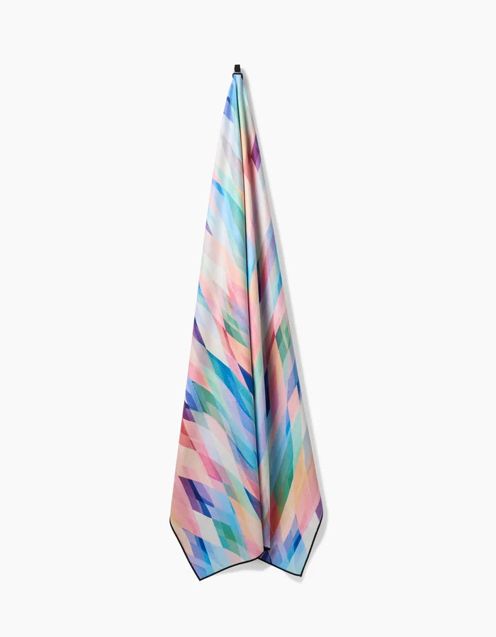 The Eventide towel by Rachel Parker is a quick-drying, recycled towel with a geometric pattern of pastel and bright diamond shapes, shown hanging from a single point against a white background.