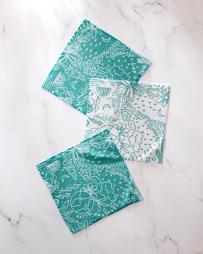 Three GEOMETRY "Every Day" cloth napkins, crafted from recycled materials, display abstract teal and white floral and doodle patterns—two with more teal, one with a mostly white design—all set on a marble surface.
