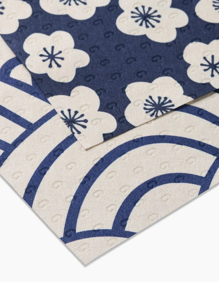 Two Every Wish sheets by GEOMETRY overlap: the top features navy blue with white plum blossoms, while the bottom shows a white base with bold blue arches and swirls. Both are reusable and textured.